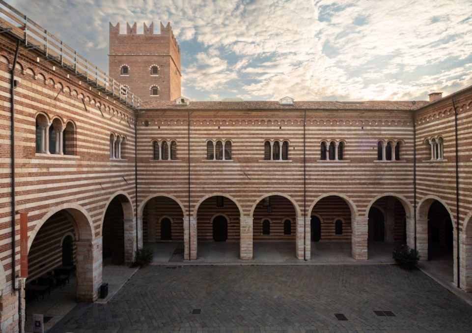 Immagine presa dall'alto del palazzo della Galleria d'Arte Moderna di Verona, si percepisce la piazza al centro del palazzo, Cortile Mercato Vecchio in basso e in alto un cielo  con un sole leggero e delle nuvole.