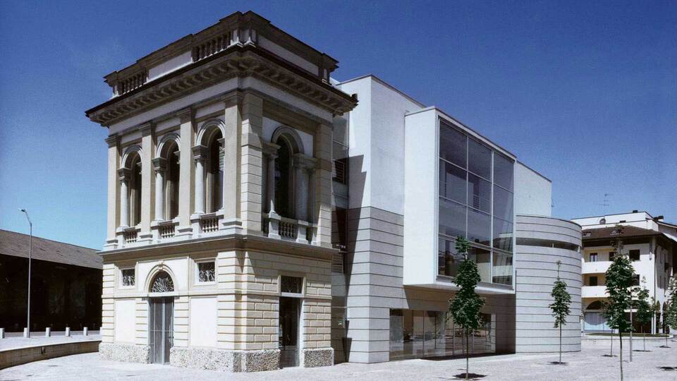 Facciata del MAC Lissone: a sinistra un edificio storico in stile neoclassico con archi e colonne, a destra un'ampia estensione moderna in vetro e pannelli bianchi, su piazza pedonale con alberi giovani.