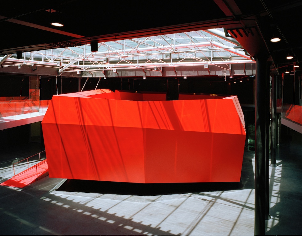 Immagine della struttura interna del MACRO, dello spazio per conferenze, simile ad una installazione poliedrica di colore rosso acceso al centro del grande ingresso, illuminata da un lucernario a struttura metallica che proietta ombre lineari sulla superficie.