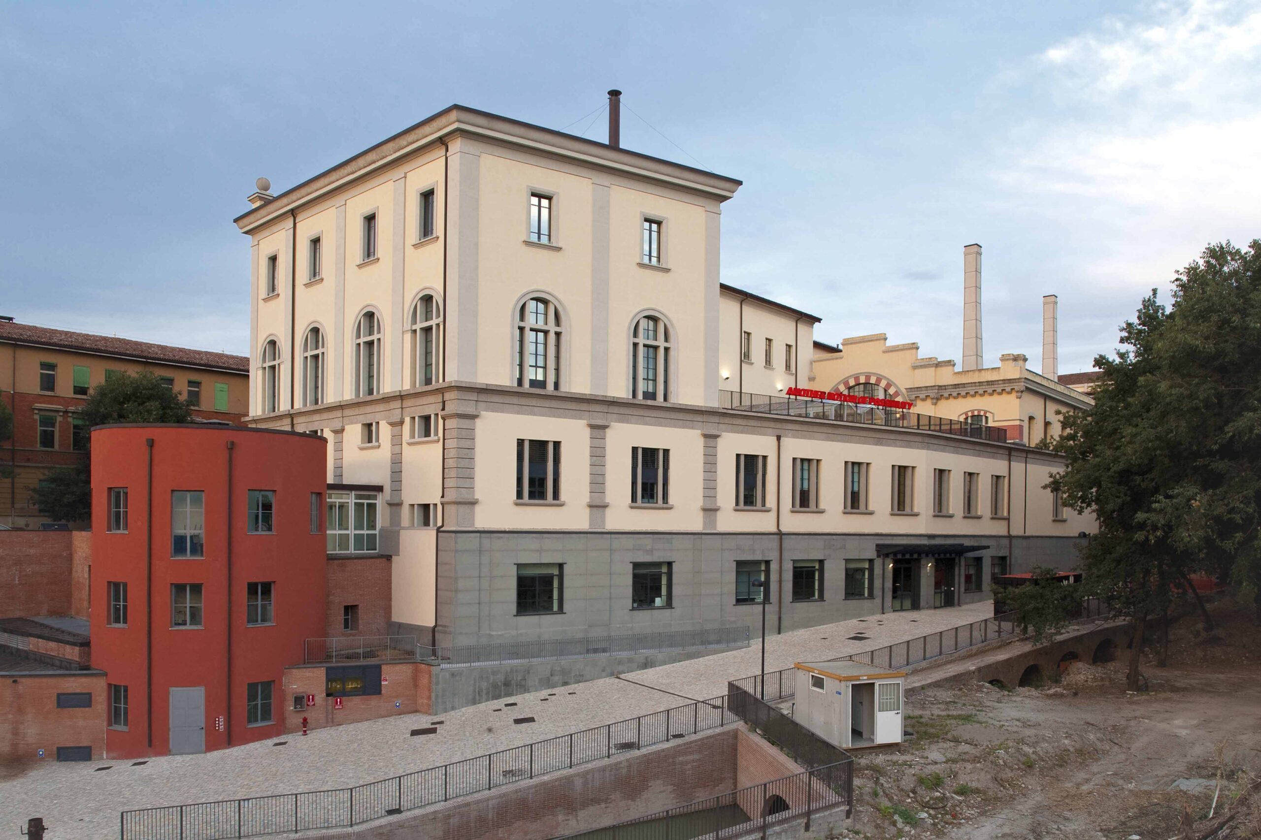 Vista esterna del MAMbo, ex complesso industriale riconvertito, con edificio principale color avorio, torre rossa in primo piano e ciminiere sullo sfondo, al crepuscolo.