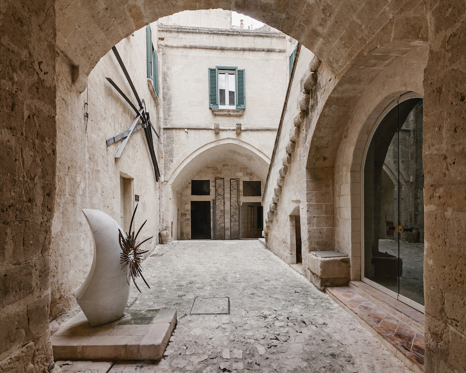 Cortile in pietra di Palazzo Pomarici, sede del MUSMA, con archi in tufo, una scultura bianca astratta in primo piano e una porta d'ingresso in ferro battuto sullo sfondo.
