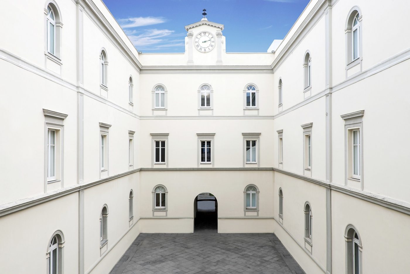 Cortile interno di Palazzo Donnaregina, con facciate bianche su tre piani, finestre ad arco simmetriche e una torre con orologio sullo sfondo di un cielo azzurro.