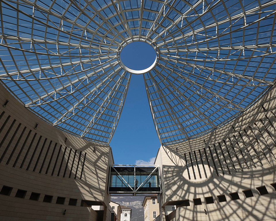 Vista dal basso della grande cupola vetrata del Mart, con struttura metallica raggiata che si apre verso il cielo azzurro. Sullo sfondo una passerella sospesa collega gli edifici del museo.
