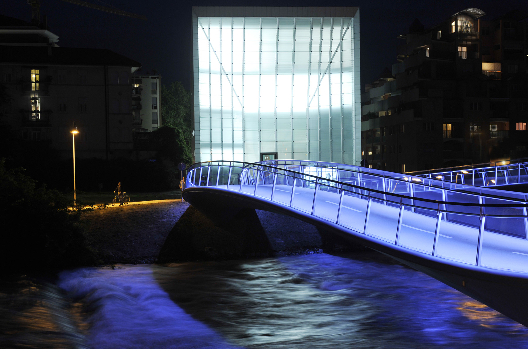 Vista notturna del Museion di Bolzano: la facciata luminosa dell'edificio si staglia sullo sfondo, mentre in primo piano un ponte pedonale illuminato di blu attraversa il fiume verso il museo.