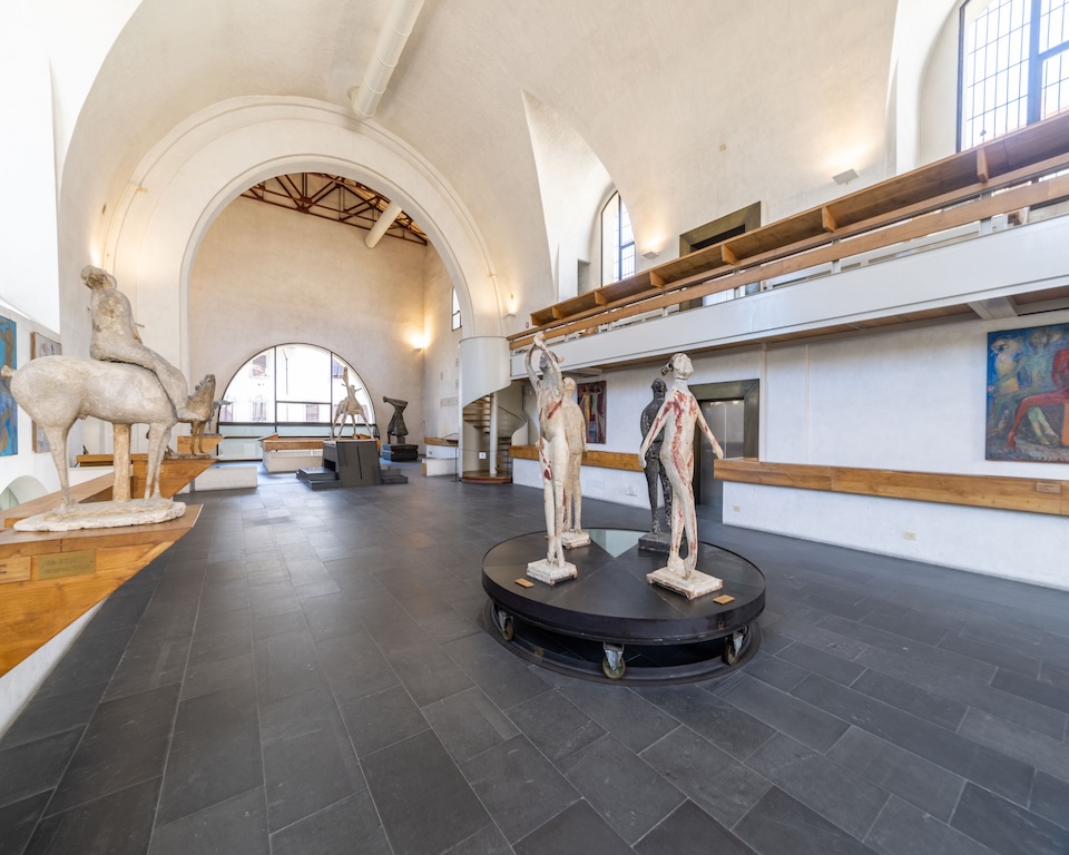 Interno del Museo Marino Marini nell'antica chiesa di San Pancrazio, con soffitto a volta bianco, sculture di cavalli e figure umane esposte su piedistalli, e dipinti alle pareti sotto un matroneo in legno.