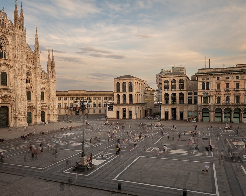 Vista al tramonto di Piazza del Duomo a Milano: sulla sinistra la facciata gotica del Duomo, sulla destra l'edificio dell'Arengario che ospita il Museo del Novecento, con la piazza animata da persone in movimento.