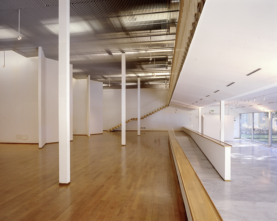 Interno del PAC di Milano: foto della sala espositiva vuota con pavimento in parquet, pareti bianche mobili, colonne bianche e una scala che conduce al livello superiore, con ampie vetrate sul fondo.