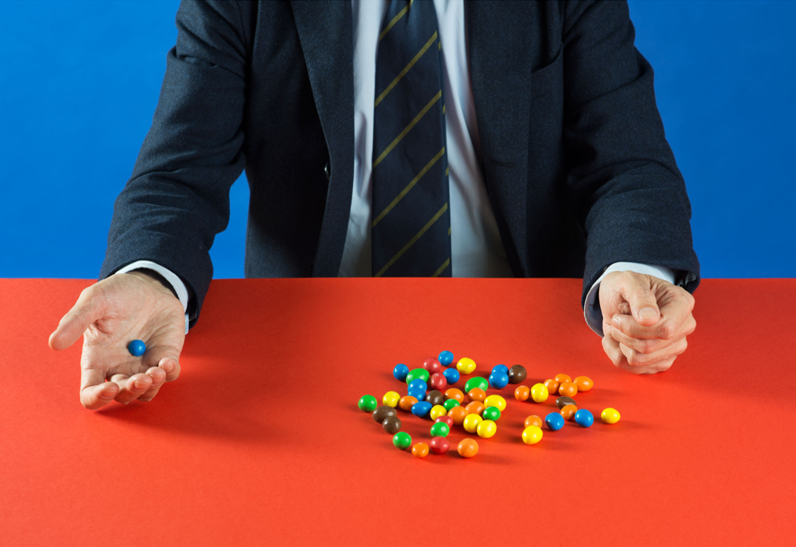 Fotografia con sfondo blu e piano rosso: un uomo in giacca e cravatta, inquadrato dal busto in giù, tiene con una mano una singola confetto colorato, mentre l'altra mano trattiene, chiusa a coppa sul tavolo, un grande mucchio di confetti colorati sparsi.